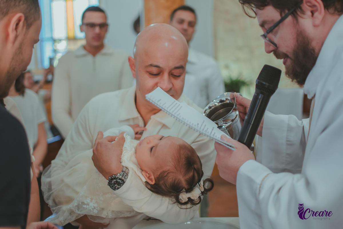 O Padre jogando água batismal na testa da bebê que está no colo de seu padrinho, durante batizado.