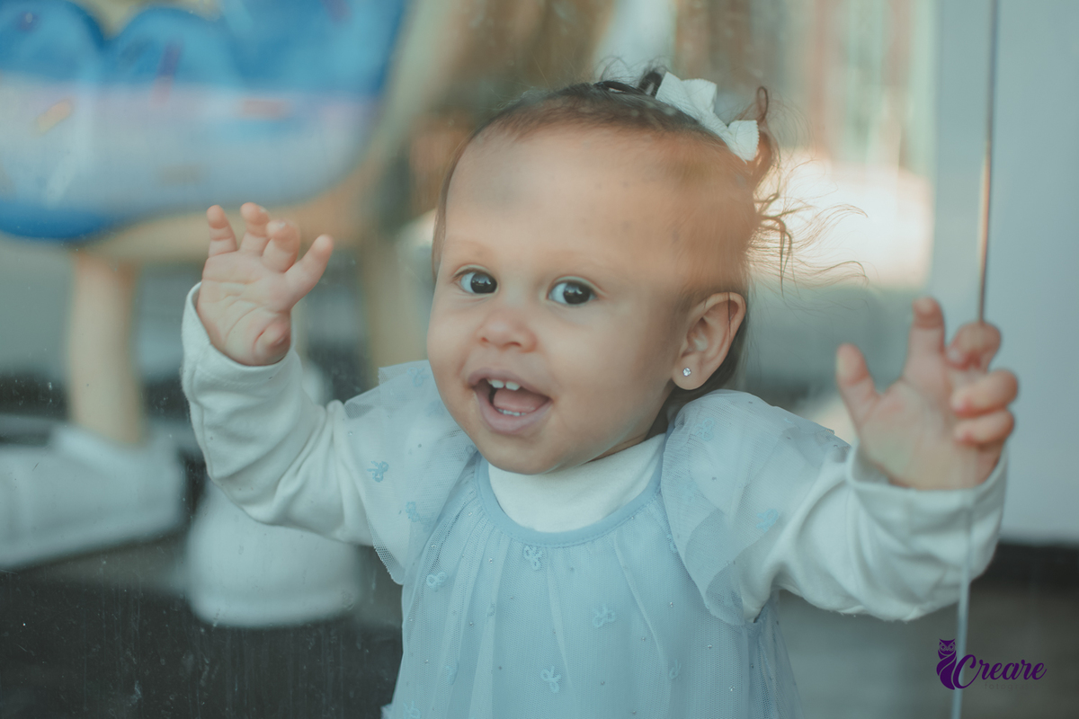 Uma bebê sorrindo em frente uma porta de vidro, durante sua festa de aniversário de 1 ano.