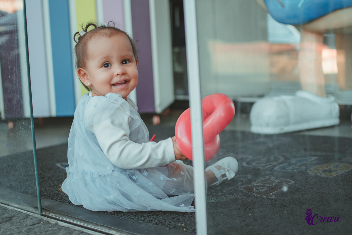Uma bebê sorrindo em frente uma porta de vidro, durante sua festa de aniversário de 1 ano.