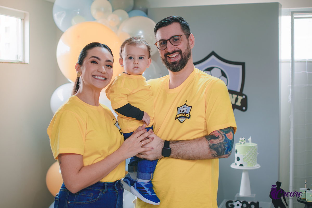Família reunida em frente à decoração de aniversário de 1 ano com tema futebol, usando camisetas amarelas e celebrando o primeiro aniversário do bebê.