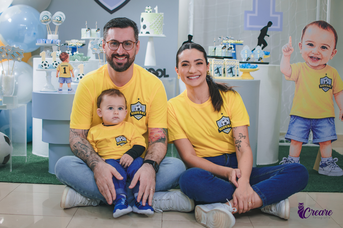 Família reunida em frente à decoração de aniversário de 1 ano com tema futebol, usando camisetas amarelas e celebrando o primeiro aniversário do bebê.