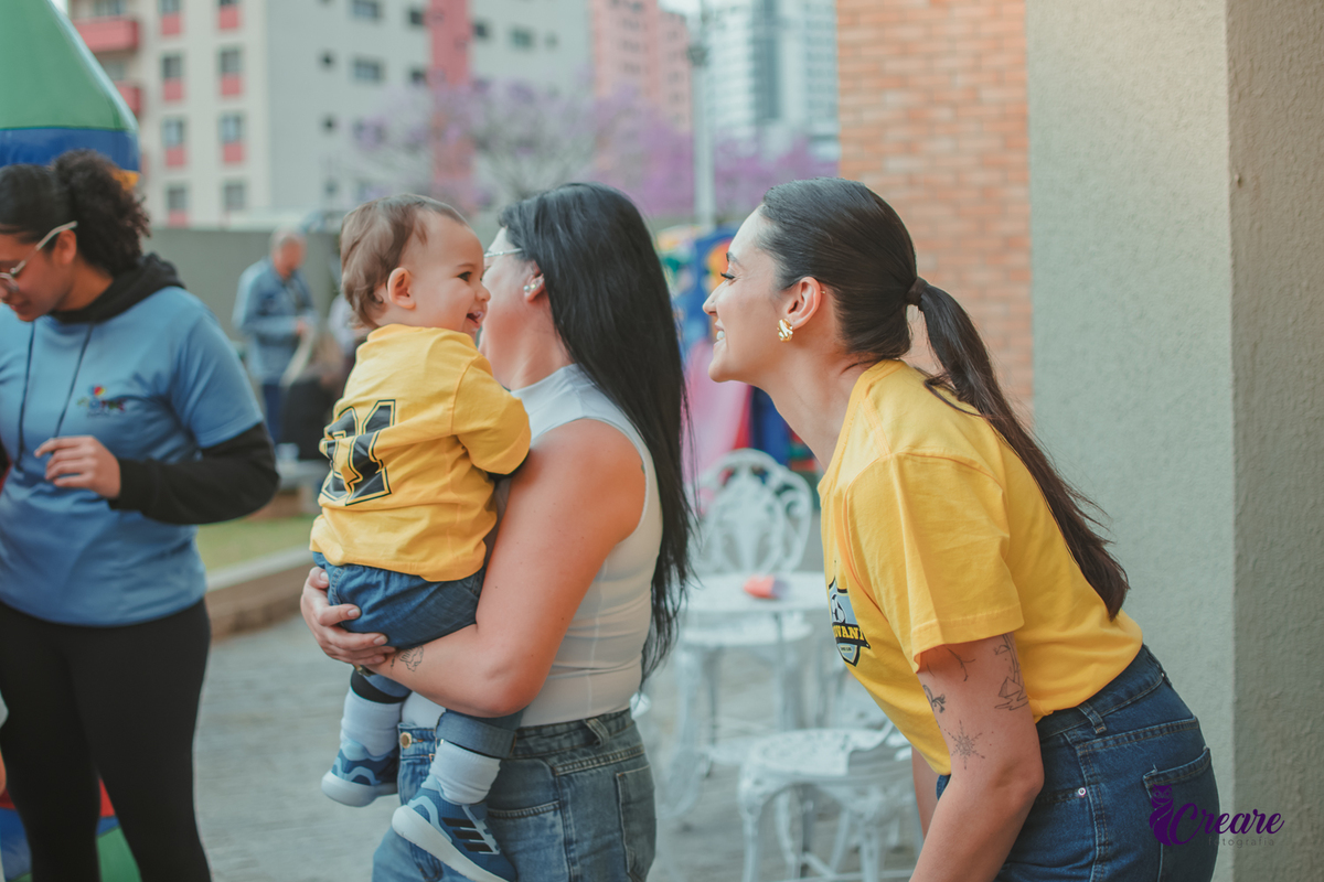 Um mulher brincando com seu bebê que está no colo de outra mulher, durante sua festa de aniversário de 1 ano.