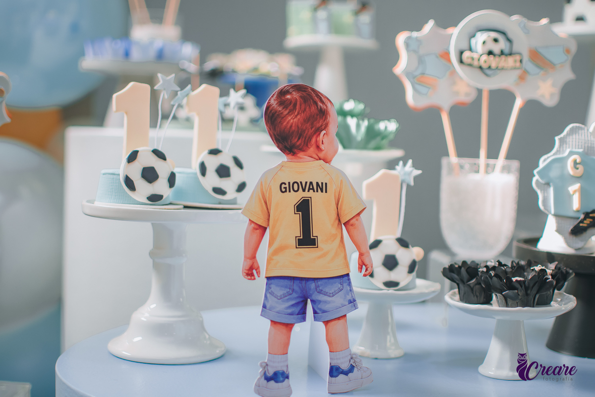 Decoração de festa de 1 ano com tema futebol, um desenho de uma criança de gostas com blusa amarela escrito Giovani e o número 1.