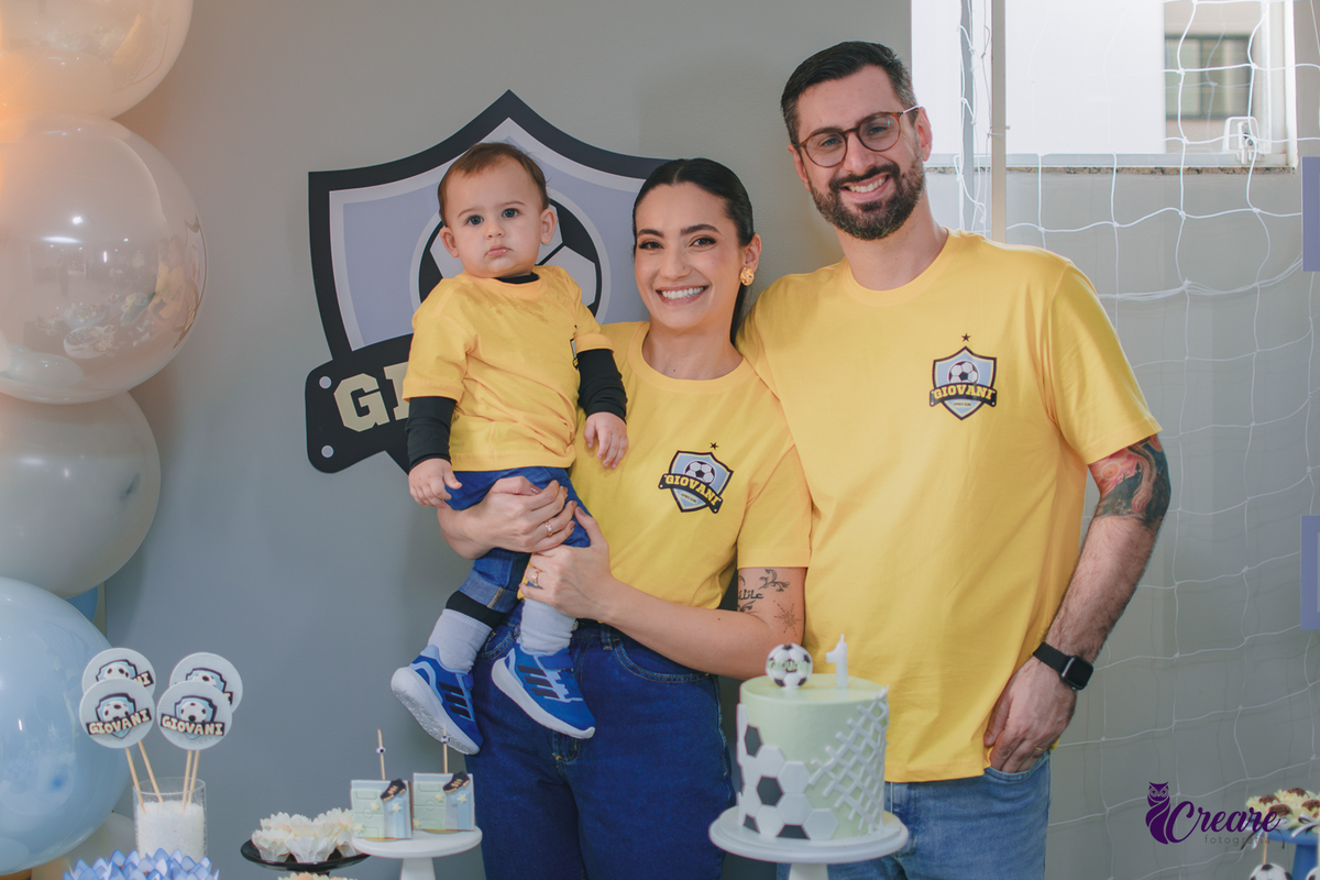 Família reunida em frente à decoração de aniversário de 1 ano com tema futebol, usando camisetas amarelas e celebrando o primeiro aniversário do bebê.