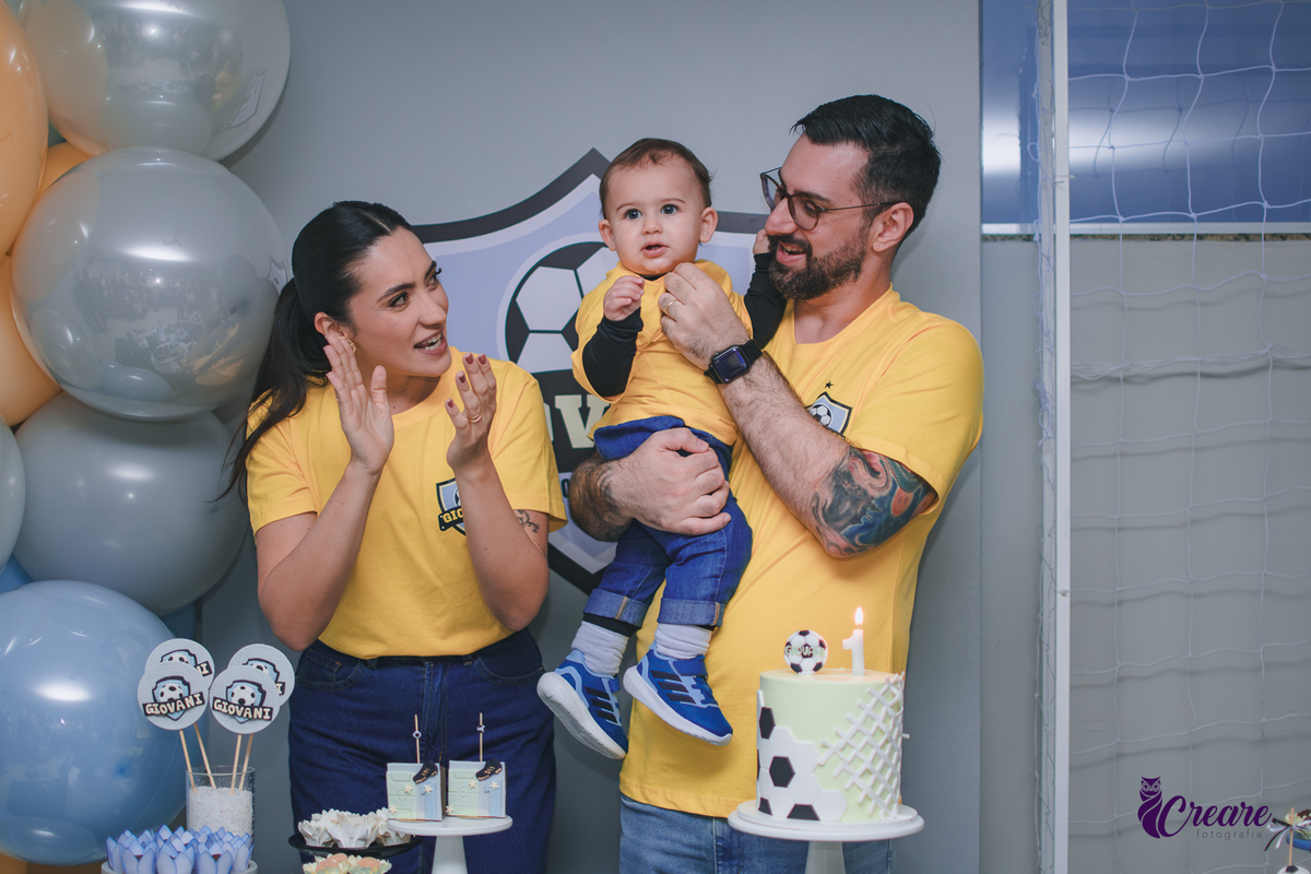Família canta parabéns para um bebê de 1 ano durante a festa de aniversário, reunida ao redor da mesa de decoração em um momento de celebração.