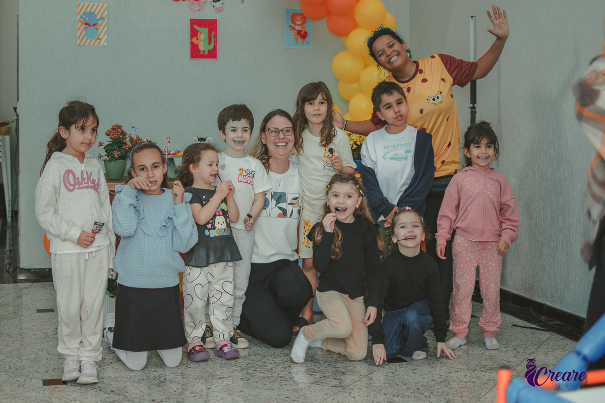 Crianças reunidas em frente mesa de decoração com balões laranjas e amarelos, durante festa de aniversário infantil em casa.