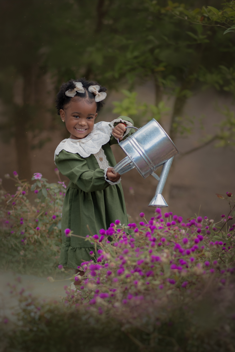 Uma criança usando vestido verde e gola branca, regando flores, com um regador de metal, durante seu ensaio fotográfico externo.