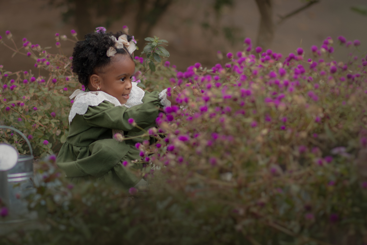 Uma criança de 2anos usando vestido verde e gola branca, entre flores roxas, durante seu ensaio fotográfico ao ar livre.
