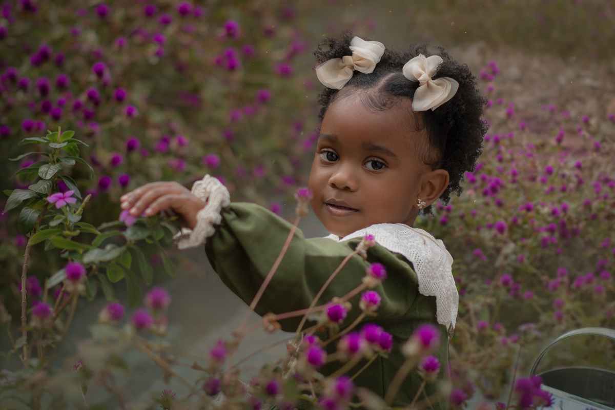 Uma criança de 2anos usando vestido verde e gola branca, entre flores roxas, durante seu ensaio fotográfico ao ar livre.