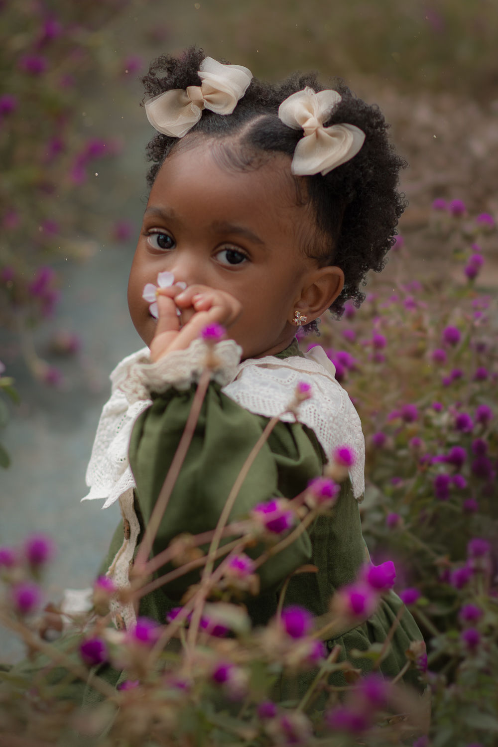 Uma criança de 2anos usando vestido verde e gola branca, cheirando uma flor rosa, durante seu ensaio fotográfico ao ar livre.