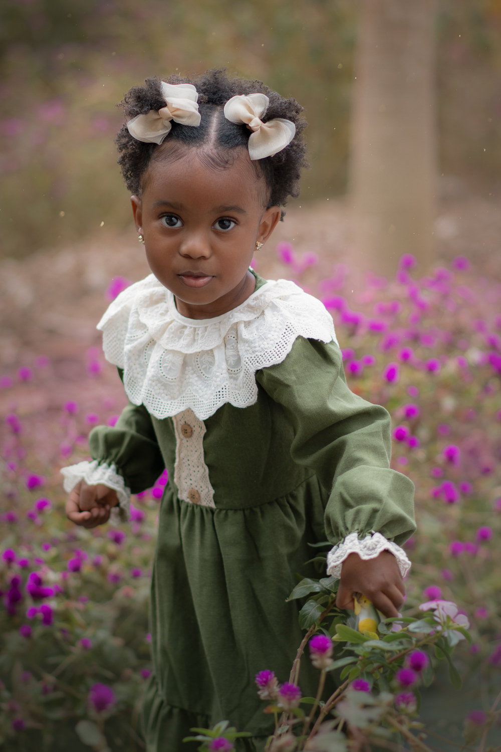Uma criança de 2anos usando vestido verde e gola branca, entre flores roxas, durante seu ensaio fotográfico ao ar livre.