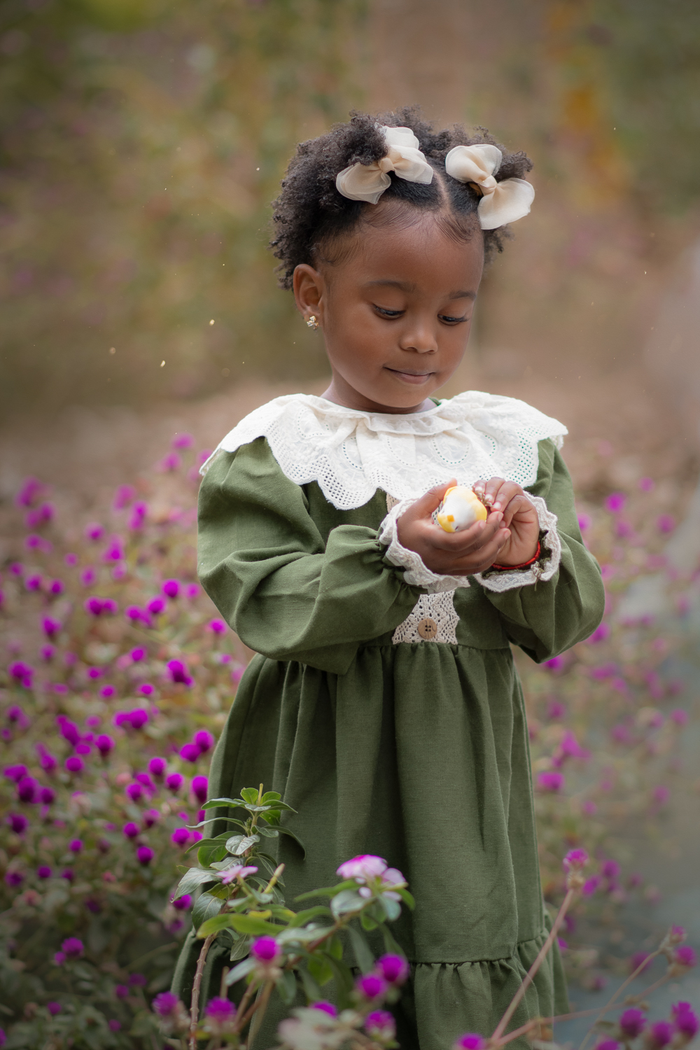 Uma criança de 2anos usando vestido verde e gola branca, entre flores roxas, durante seu ensaio fotográfico ao ar livre.