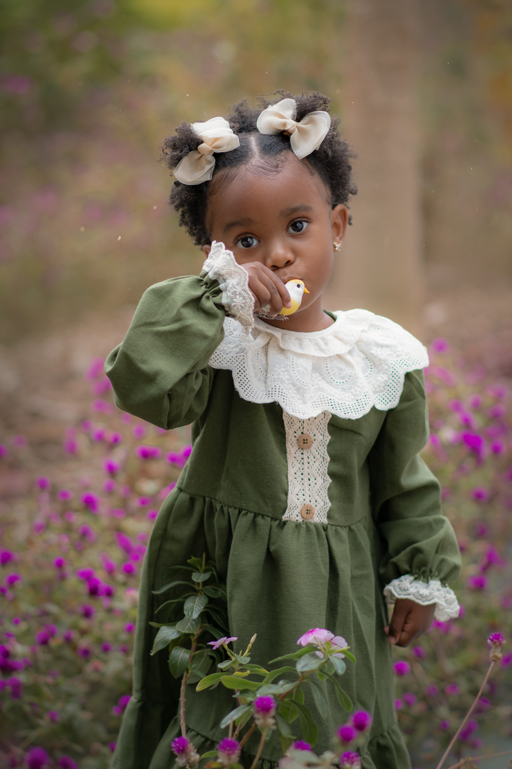 Uma criança de 2anos usando vestido verde e gola branca, entre flores roxas, durante seu ensaio fotográfico ao ar livre.