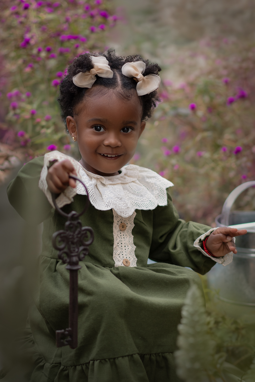 Uma criança de 2anos usando vestido verde e gola branca, segurando uma chave de ferro antiga e sorrindo, durante seu ensaio fotográfico ao ar livre.