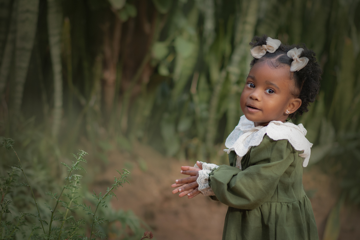 Uma criança negra, usando vestido verde e olhando para frente com uam sorriso, durante seu ensaio fotográfico externo.