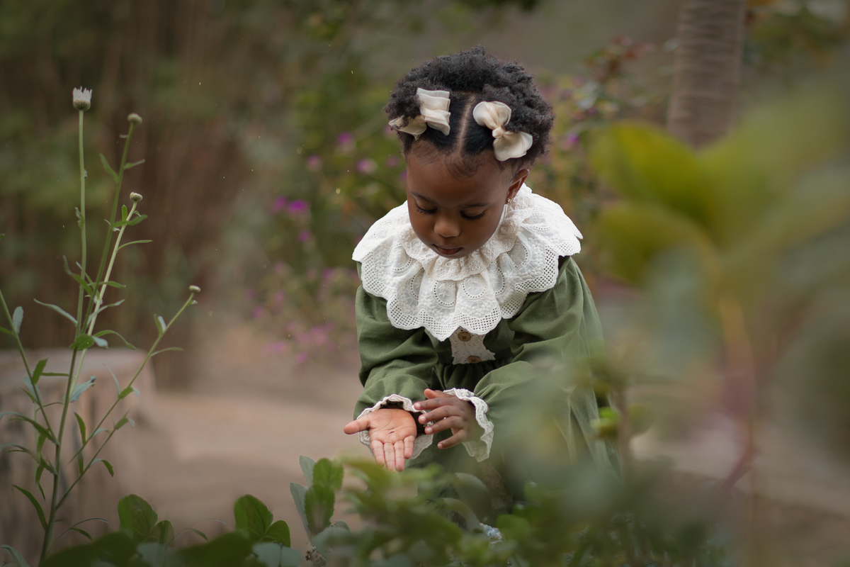 Uma criança usando vestido verde e gola branca, plantando semente de flores, durante seu ensaio fotográfico externo.