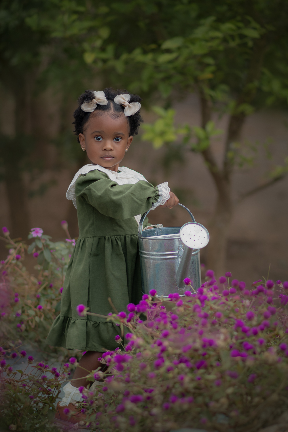 Uma criança usando vestido verde e gola branca, regando flores, com um regador de metal, durante seu ensaio fotográfico externo.