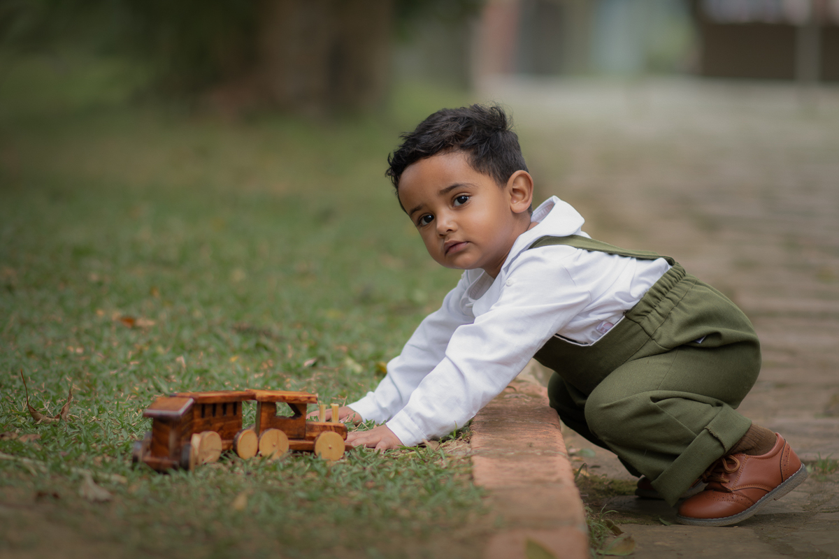 Um menino usando macacão verde e brincando na grama com um trenzinho de madeira, durante seu ensaio fotográfico em Ribeirão Pires.