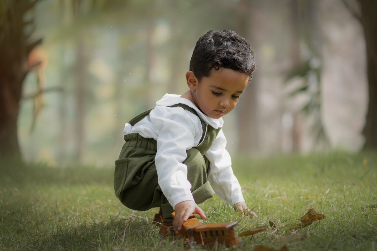 Um menino usando macacão verde e brincando na grama com um trenzinho de madeira, durante seu ensaio fotográfico em Ribeirão Pires.
