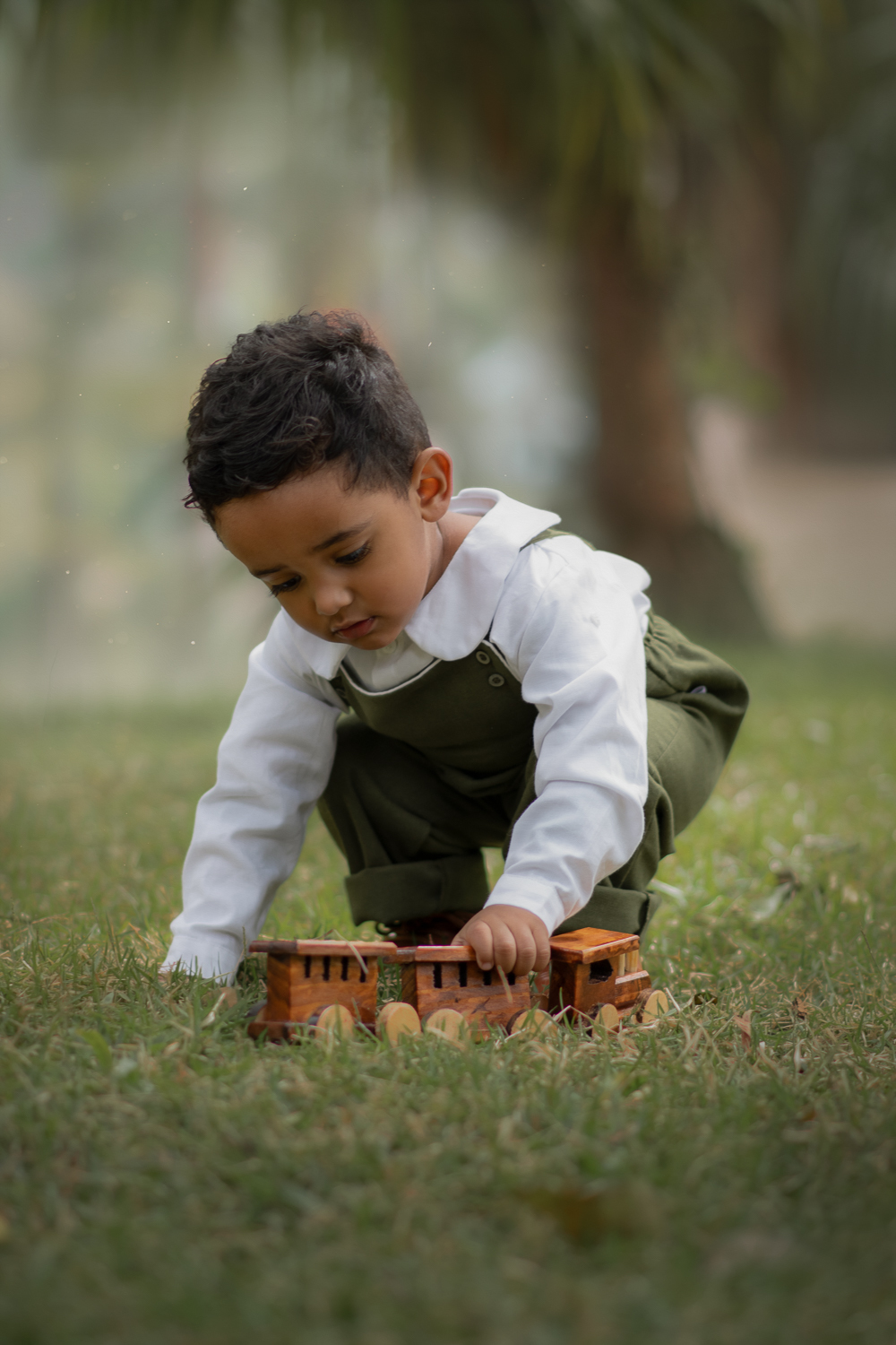 Um menino usando macacão verde e brincando na grama com um trenzinho de madeira, durante seu ensaio fotográfico em Ribeirão Pires.