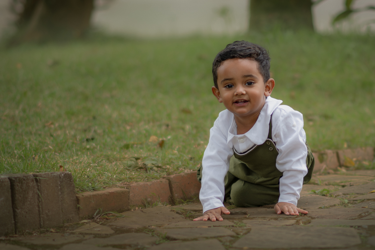 Um menino de 2 anos, engatinhando na grama e sorrindo, durante seu ensaio fotográfico no Parque Luis Carlos Grecco.