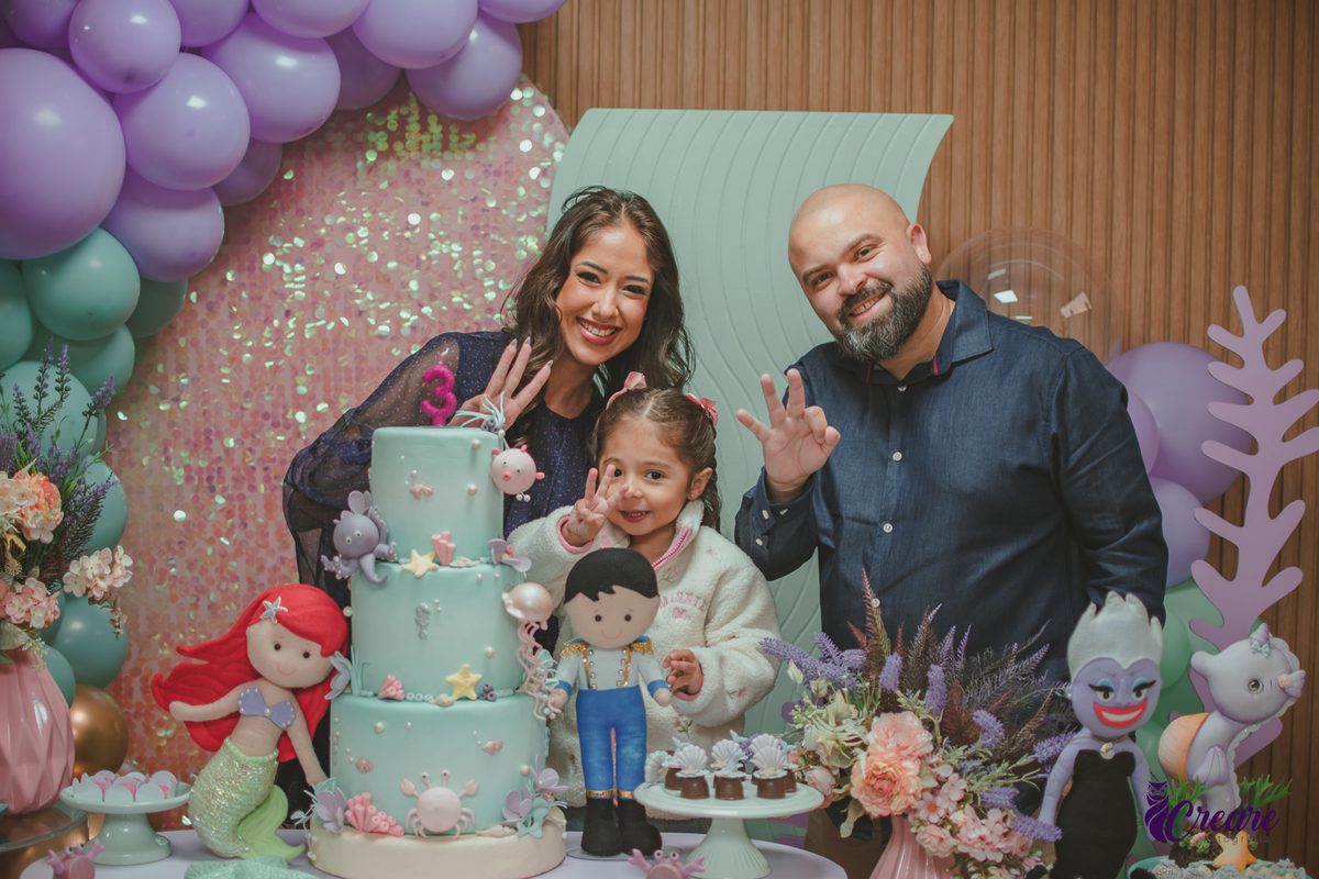 Um homem, com sua esposa e sua filha, atrás de uma mesa de decoração de festa de aniversário com tema A pequena sereia.