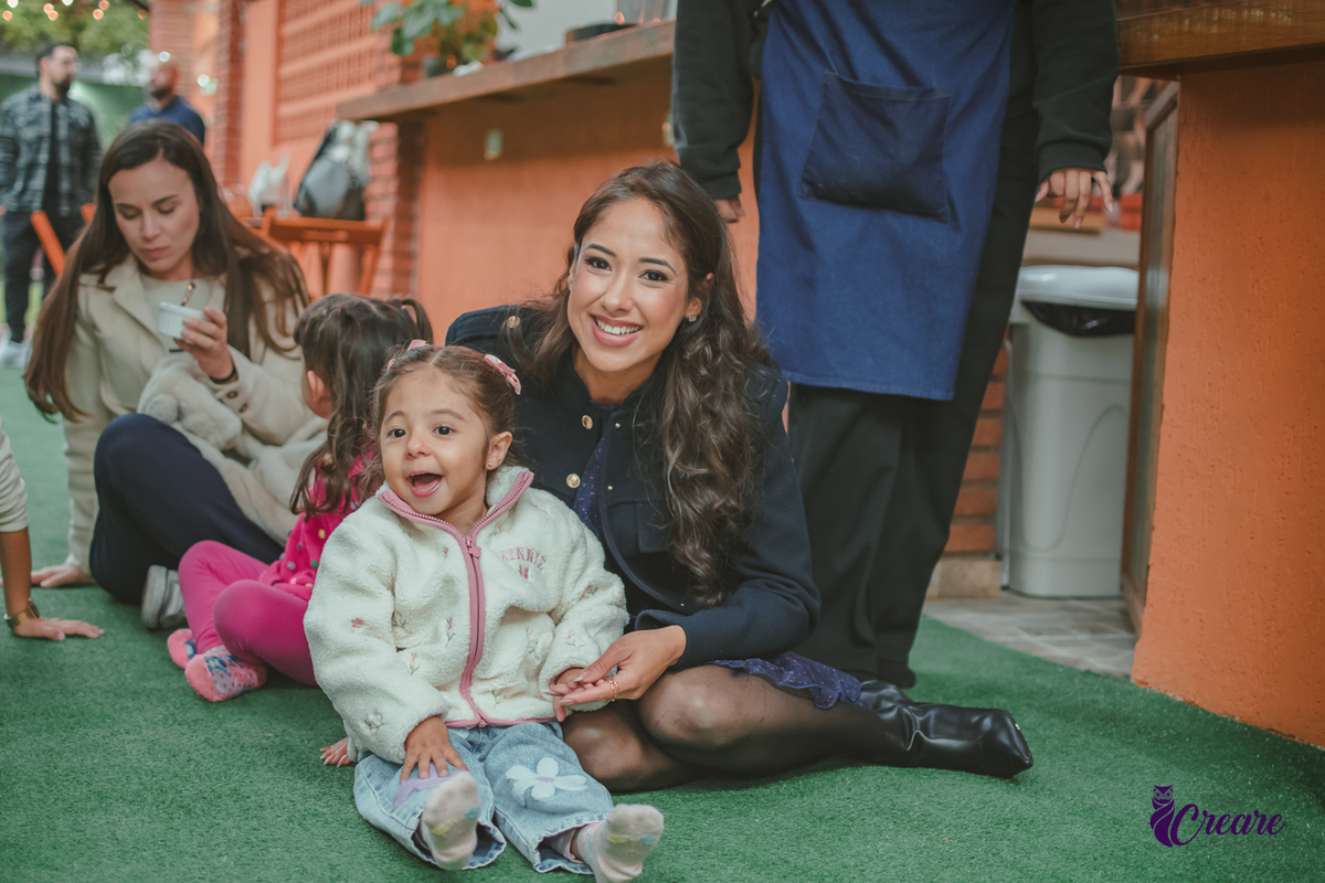 Uma mãe de sua filha sentanda na grama do buffet Patuska Brincadeiras, durante apresentação no buffet Patuska Brincadeiras.