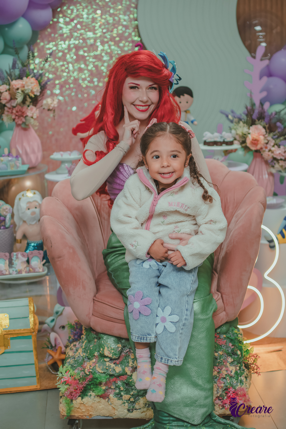 Uma criança, sentada no colo da Personagem Ariel, da Pequena Sereia, durante sua festa de aniversário infantil.