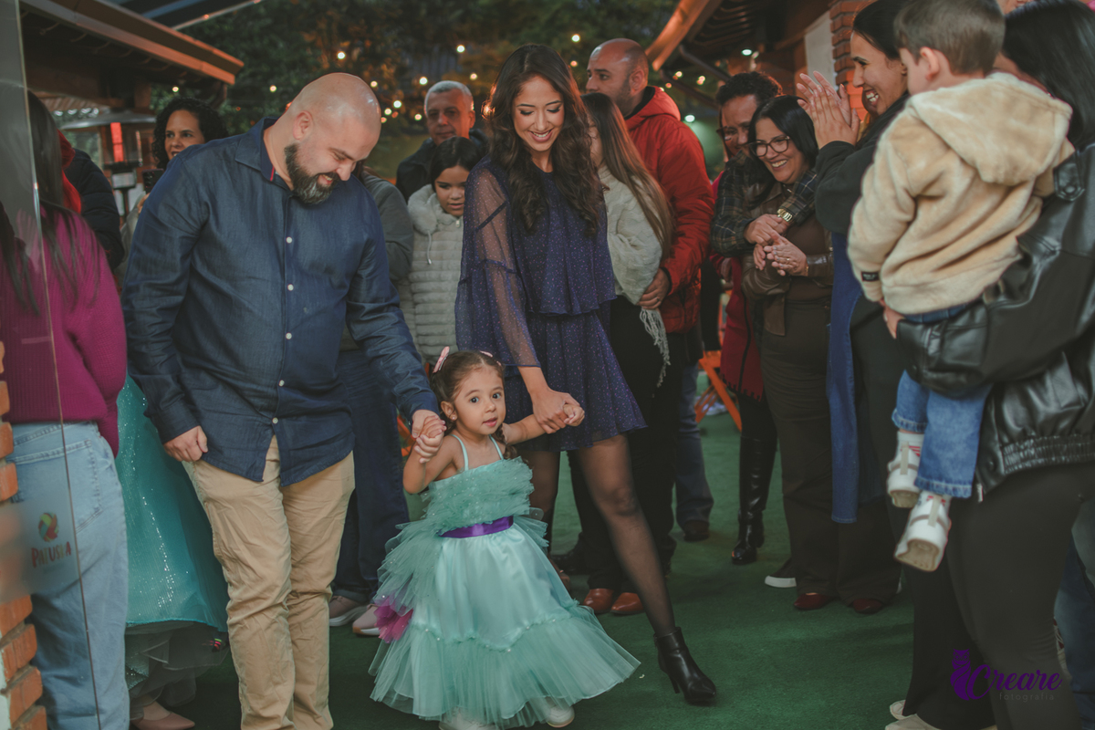 Uma família entrando em uma festa com a criança usando um vestido verde água, pessoas felizes ao redor.