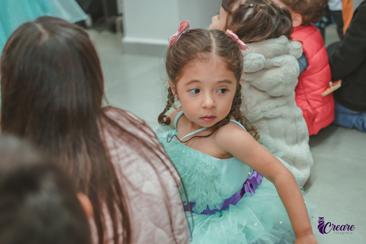 Uma menina usando vestido Verde, sentada no chão com outras crianças em volta, durante festa de aniversário.