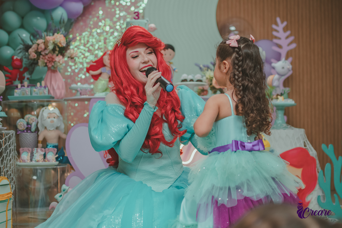 Personagem Ariel, cantando para uma criança, que usa um vestido verde igual ao da personagem, durante festa de aniversário.
