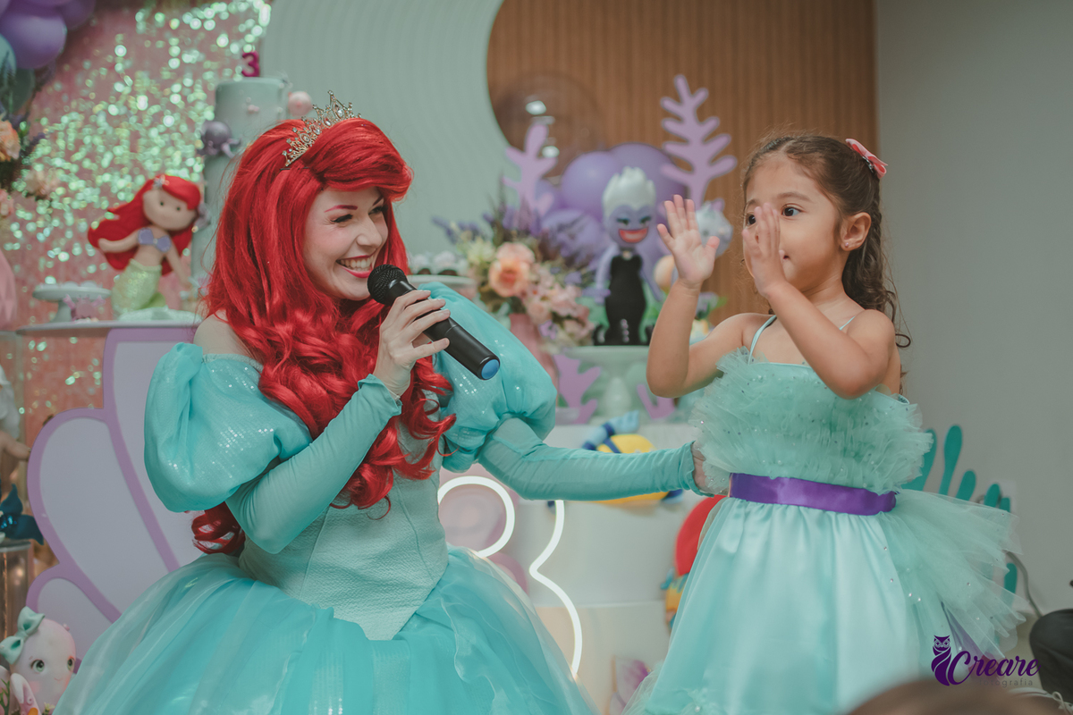 Personagem Ariel, cantando para uma criança, que usa um vestido verde igual ao da personagem, durante festa de aniversário.