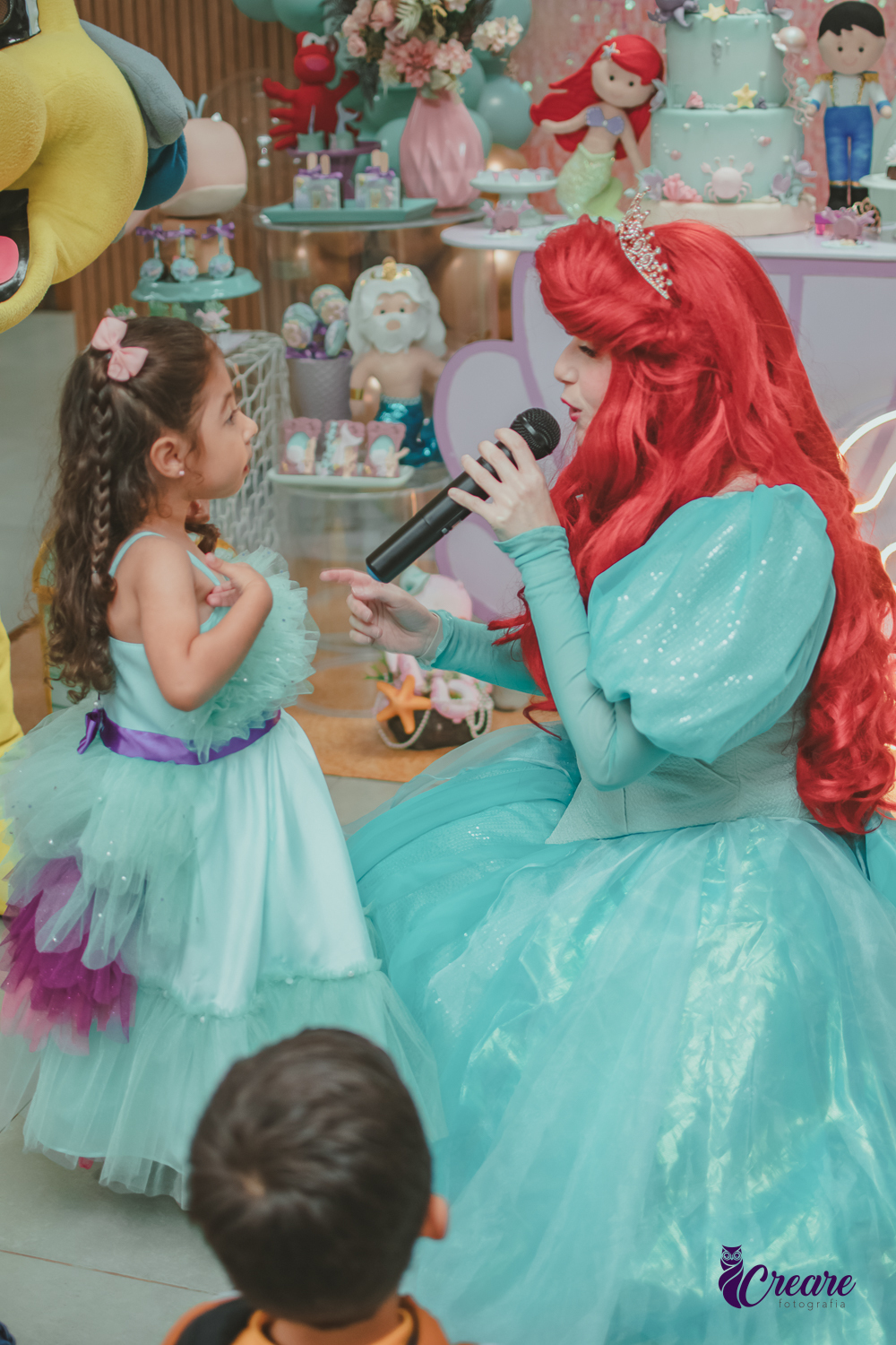 Personagem Ariel, cantando para uma criança, que usa um vestido verde igual ao da personagem, durante festa de aniversário.