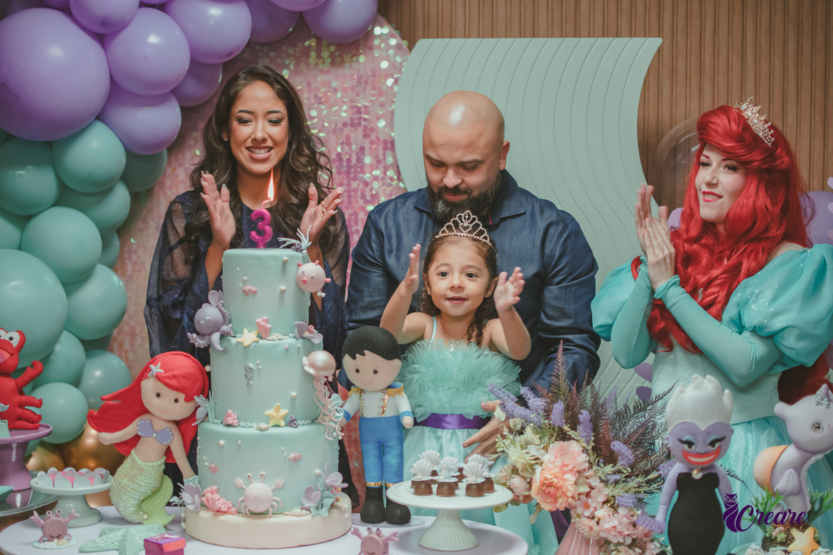 uma família e a personagem Ariel, cantando parabéns para uma criança, durante sua festa de aniversário.