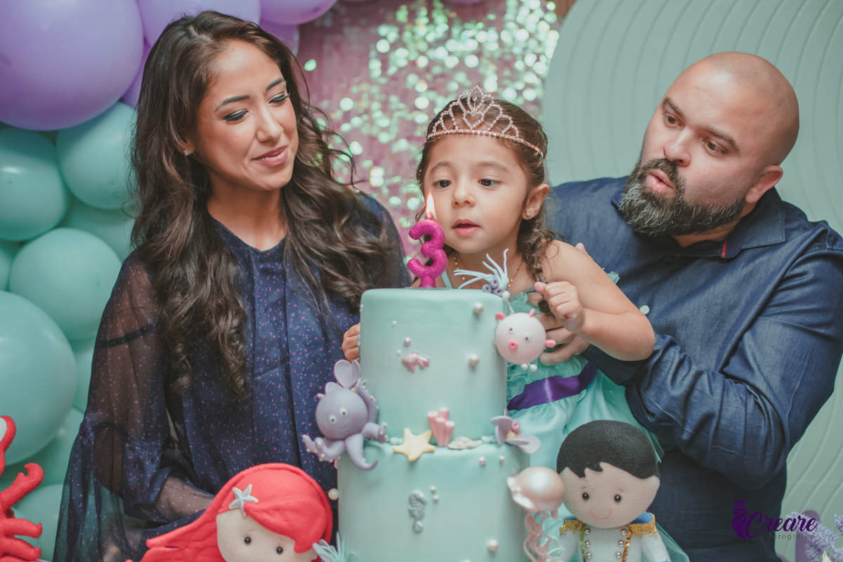 Uma criança assoprando a velinha do seu aniversário, ao lado da sua mãe e seu pai.