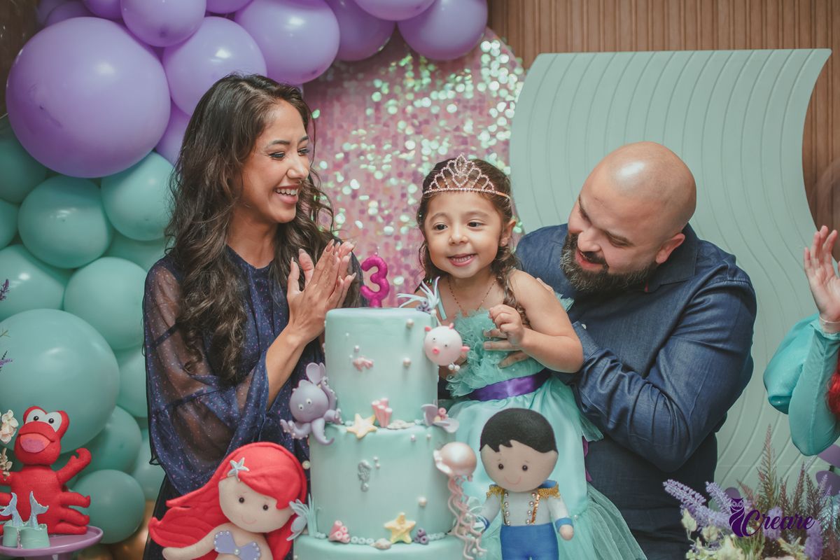 Uma criança assoprando a velinha do seu aniversário, ao lado da sua mãe e seu pai.