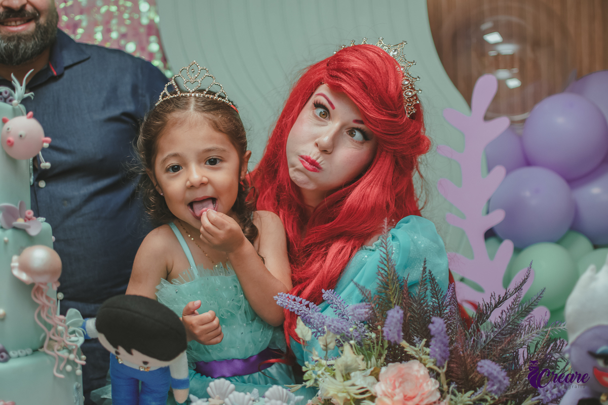 Uma criança fazendo careta, junto com a personagem Ariel, da Pequena Sereia, durante festa de aniversário.