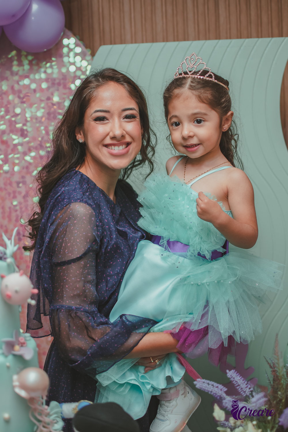 Uma mulher segurando no colo sua filha, que usa um vestido verde, durante festa de aniversário infantil.