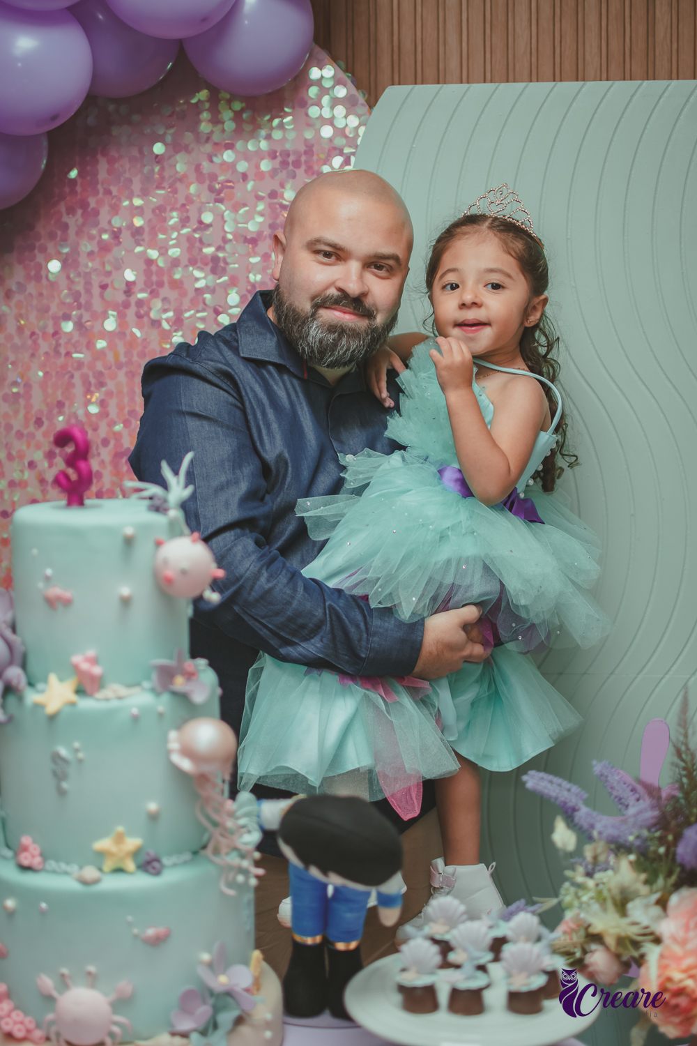 Um homem segurando no colo sua filha, que usa um vestido verde, durante festa de aniversário infantil.