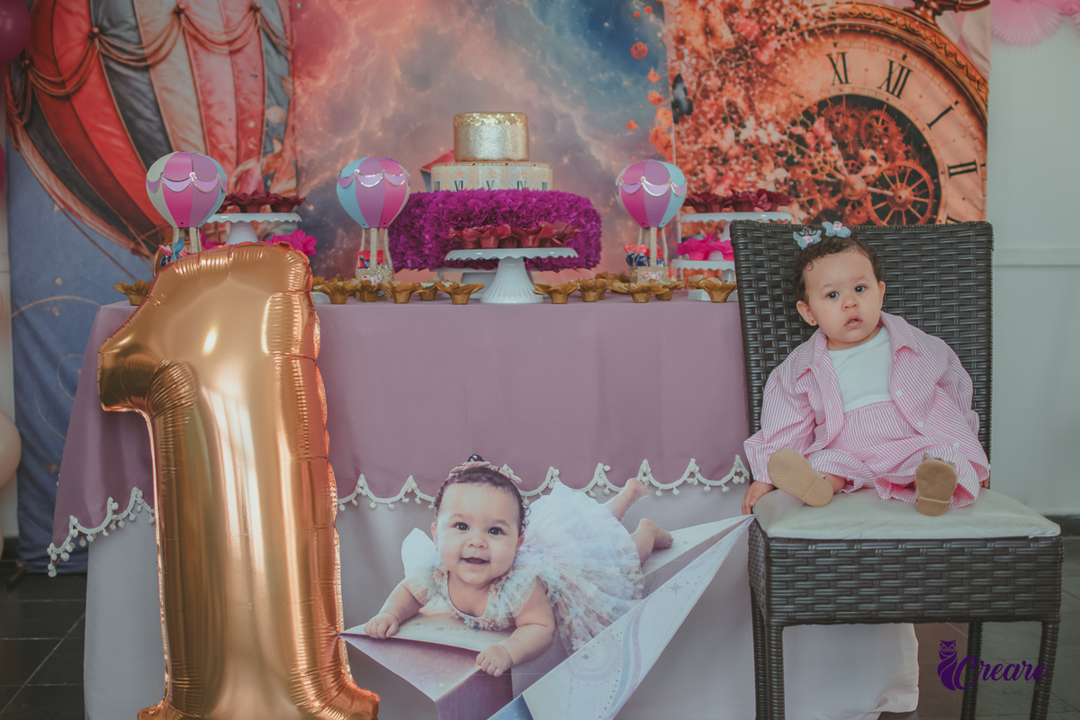 Uma menina sentada em um cadeira em frente a mesa decorada para seu aniversário de 1 ano.