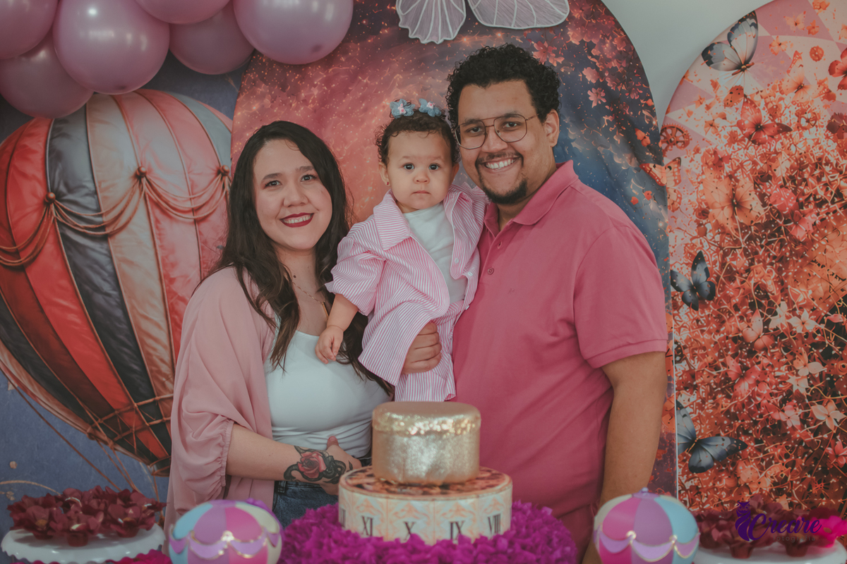 Um casal com uma bebê em frente uma mesa decorada para o aniversário de 1 ano em casa.