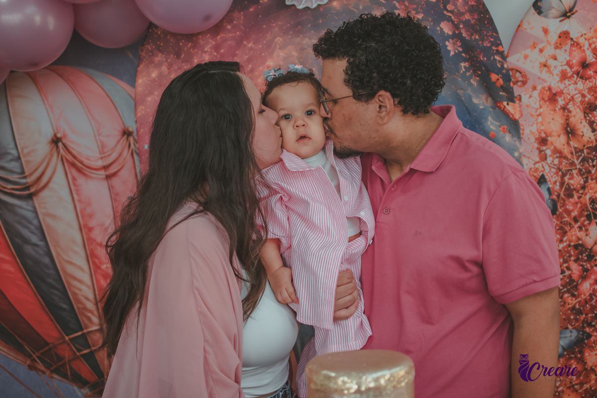 Um casal beijando o rosto de uma bebê de 1 ano, durante festa de aniversário em casa.