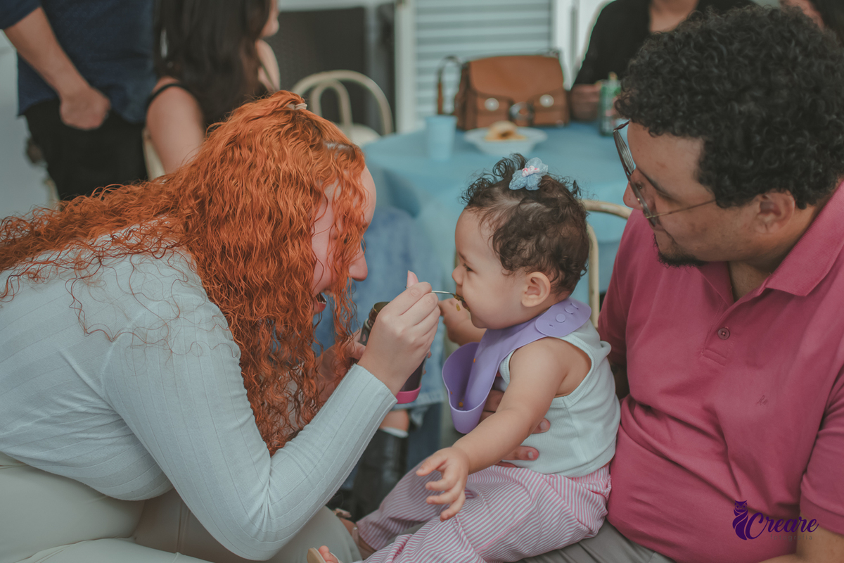 Uma mulher dando comida na boca de uma bebê que está no colo do pai, durante festa de aniversário de 1 ano em casa.