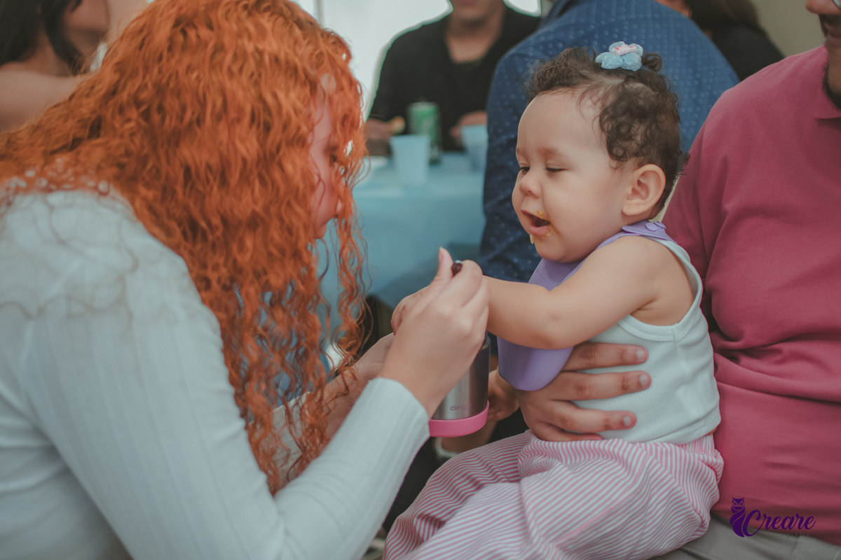 Uma mulher dando comida na boca de uma bebê que está no colo do pai, durante festa de aniversário de 1 ano em casa.