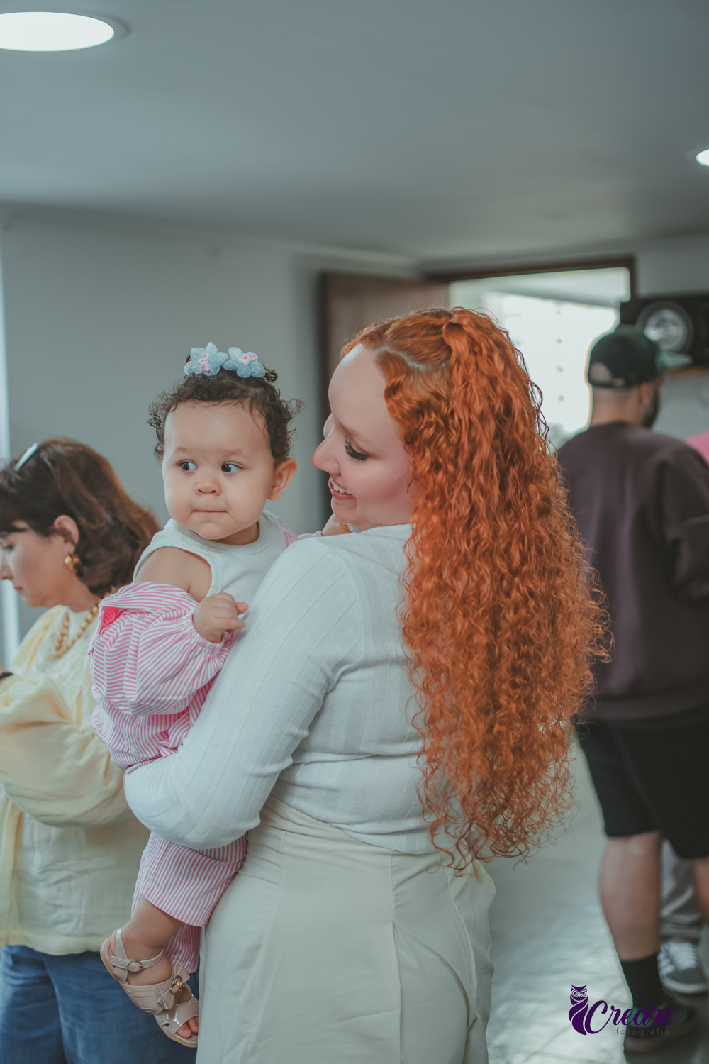 Uma mulher usando roupa branca, segurando uma bebê no colo, durante festa de aniversário de 1 ano em casa.