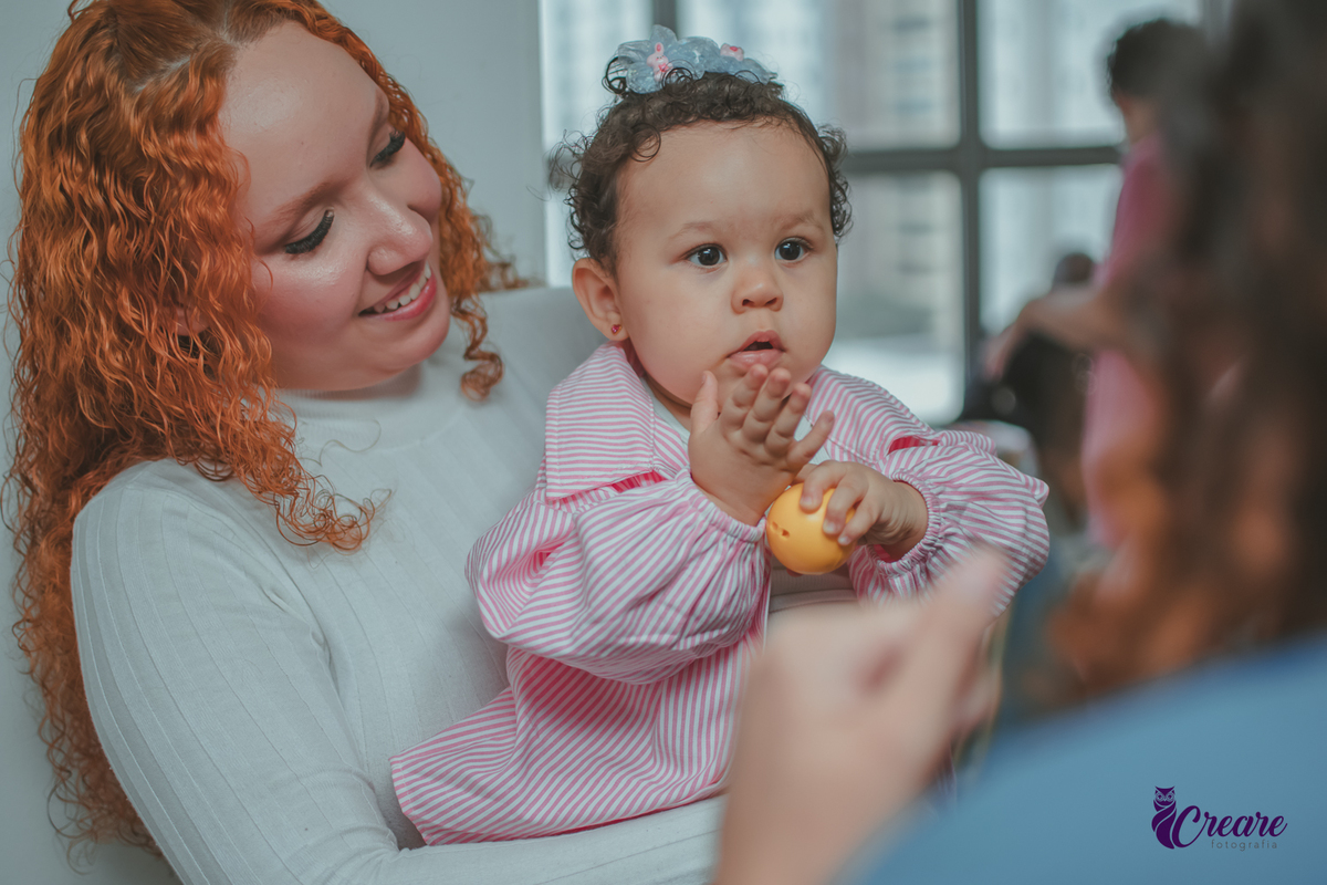 Uma mulher usando roupa branca, segurando uma bebê no colo, enquanto ela brinca com uma bolinha amarela, durante festa de aniversário de 1 ano em casa.