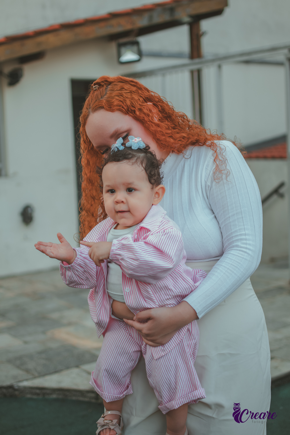 Uma bebê, apontando para sua mão, enquanto brinca no colo de uma mulher de branco, durante sua festa de aniversário de 1 ano.