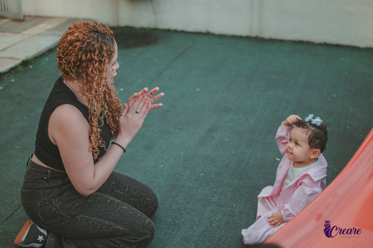 Uma mulher brincando de bater palma com uma bebê, no parquinho do condomínio, durante festa de aniversário em casa.