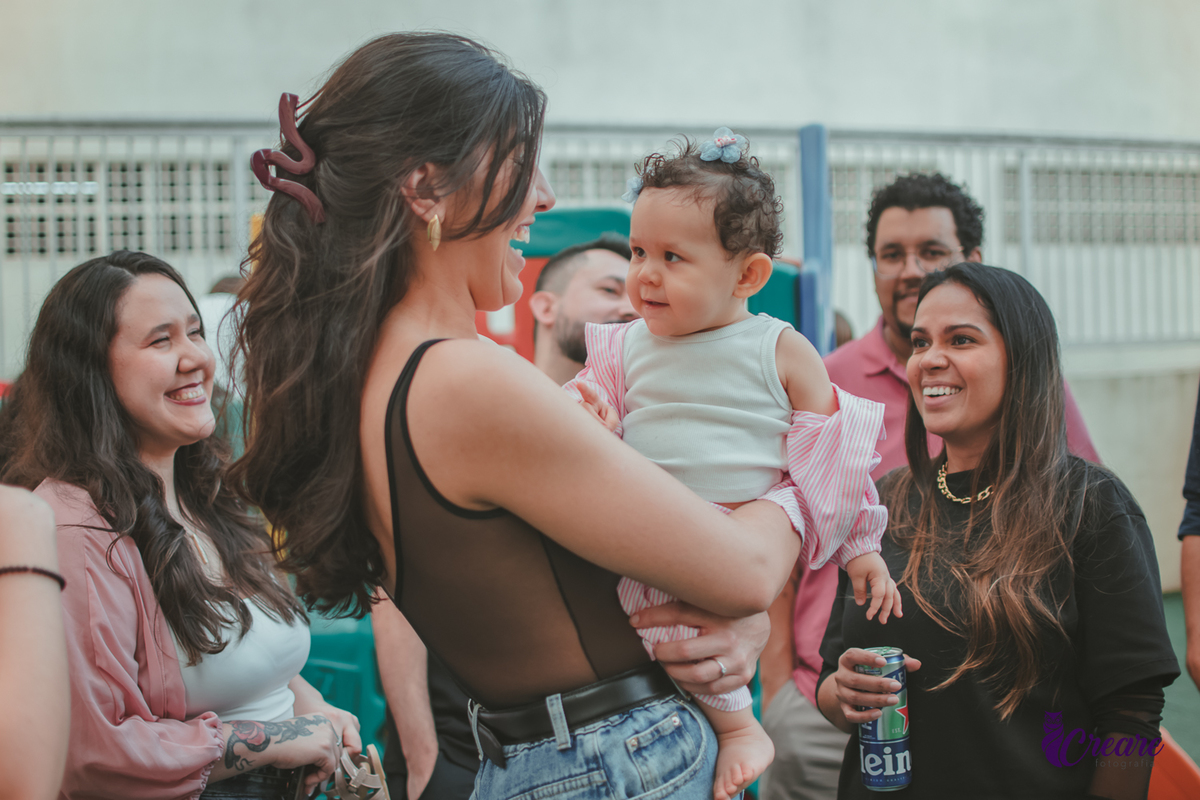 Uma mulher sorrindo com uma bebê no colo e várias pessoas ao lado sorrindo, durante festa de aniversário em casa.