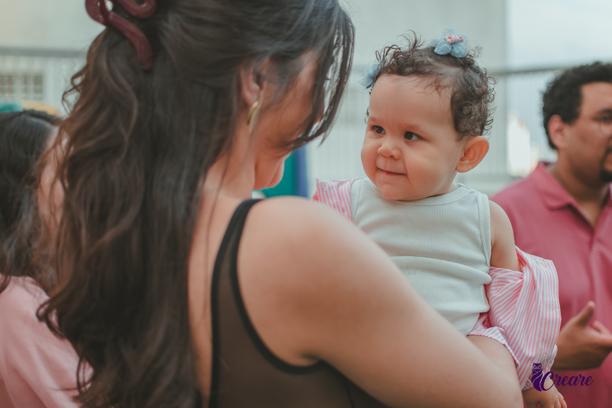 Uma mulher sorrindo com uma bebê no colo e várias pessoas ao lado sorrindo, durante festa de aniversário em casa.